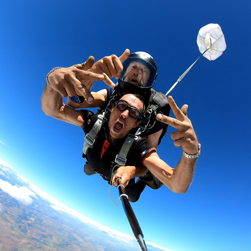 Duo de parachutistes en chute libre faisant des signes de victoire, avec un parachute principal déployé en arrière-plan